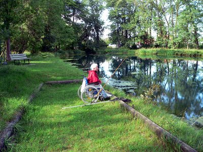 Pfaffenwinkelweiher mit Bäumen, eine Rampe führt zum Wasser, ein Angler im Rollstuhl versucht sein Glück.