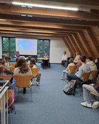In einem großen Raum mit Holzbalken-Decke stehen Stuhlreihen und vorn im Rollstuhl sitzend vor einer Leinwand Raul Krauthausen. Er trägt eine graue Schiebermütze und ein kariertes Hemd. Auf der Leinwand sieht man das Titelbild seines Buches. Ein gezeichnetes Mädchen mit einem weißen Raumfahreranzug und entsprechendem Helm. Im Hintergrund sieht man den Mond und Planeten.