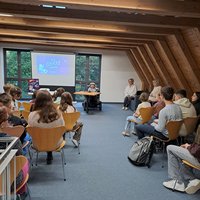 In einem großen Raum mit Holzbalken-Decke stehen Stuhlreihen und vorn im Rollstuhl sitzend vor einer Leinwand Raul Krauthausen. Er trägt eine graue Schiebermütze und ein kariertes Hemd. Auf der Leinwand sieht man das Titelbild seines Buches. Ein gezeichnetes Mädchen mit einem weißen Raumfahreranzug und entsprechendem Helm. Im Hintergrund sieht man den Mond und Planeten.