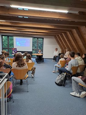 In einem großen Raum mit Holzbalken-Decke stehen Stuhlreihen und vorn im Rollstuhl sitzend vor einer Leinwand Raul Krauthausen. Er trägt eine graue Schiebermütze und ein kariertes Hemd. Auf der Leinwand sieht man das Titelbild seines Buches. Ein gezeichnetes Mädchen mit einem weißen Raumfahreranzug und entsprechendem Helm. Im Hintergrund sieht man den Mond und Planeten.