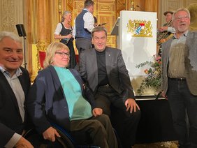 Auf dem Foto sitzt vor der Bühne Harald Güller neben Frau Gotzes. In der Mitte sitzt Ministerpräsident Markus Söder. Rechts steht Herman Wetzel aus Memmingen. Im Hintergrund sieht man Musikanten in Tracht, die am Abend auftraten. 
