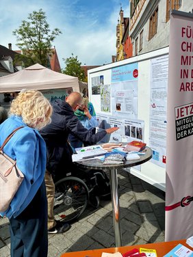 Vor einer Stellwand mit Informationen und Quizfragen sitzt ein Herr im Rollstuhl und liest aufmerksam. Hinter ihm stehen ein Mann und eine Frau, die ebenfalls die Stellwand aufmerksam Betrachten. 