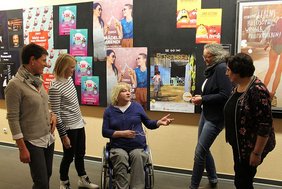 Die Personen stehen vor einer Wand mit verschiednen Kinoplakaten. Eine Person sitzt im Rollstuhl. Im Hintergrund ist das Poster zum Film "Menschsein" zu sehen. 