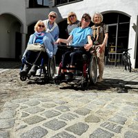 Im Vordergrund sitzen Verena Gotzes und Natalie im Rollstuhl, dahinter stehen Inge Oberst, Simone Magel-Dipre, sowie Inrid Willner-Sambs. Vorn im Bild sieht man das Kopfsteinpflaster mit der neuen, hellen Verfugung. Auf Grund des guten Wetters tragen die Teilnehmenden Sonnenbrillen. 