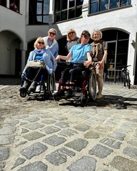 Im Vordergrund sitzen Verena Gotzes und Natalie im Rollstuhl, dahinter stehen Inge Oberst, Simone Magel-Dipre, sowie Inrid Willner-Sambs. Vorn im Bild sieht man das Kopfsteinpflaster mit der neuen, hellen Verfugung. Auf Grund des guten Wetters tragen die Teilnehmenden Sonnenbrillen. 